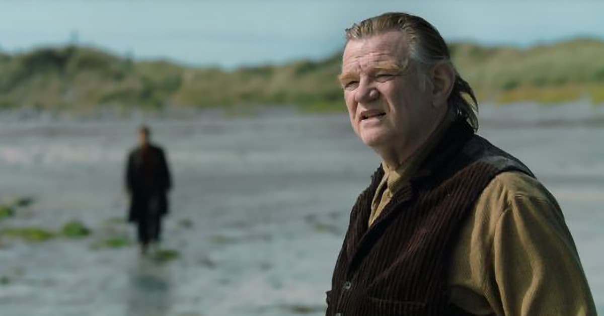 A weathered man in a corduroy vest stands on a rocky shoreline, his expression tense, as a lone figure walks away in the blurred background.