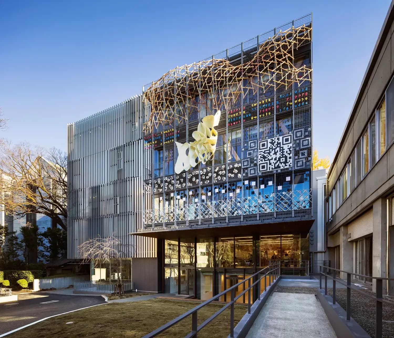 A modern and highly artistic exterior view of the Tokyo University of the Arts (Geidai) building. The right side of the glass facade features a large integrated QR code, a prominent, wavy abstract white sculpture hanging on the exterior wall, and an intricate, lattice-like wooden installation running along the roofline. The left side of the building is clad in sleek vertical metal louvers. In the foreground, a concrete ramp with grey metal railings leads down toward the entrance next to a small lawn under a clear, bright blue sky.