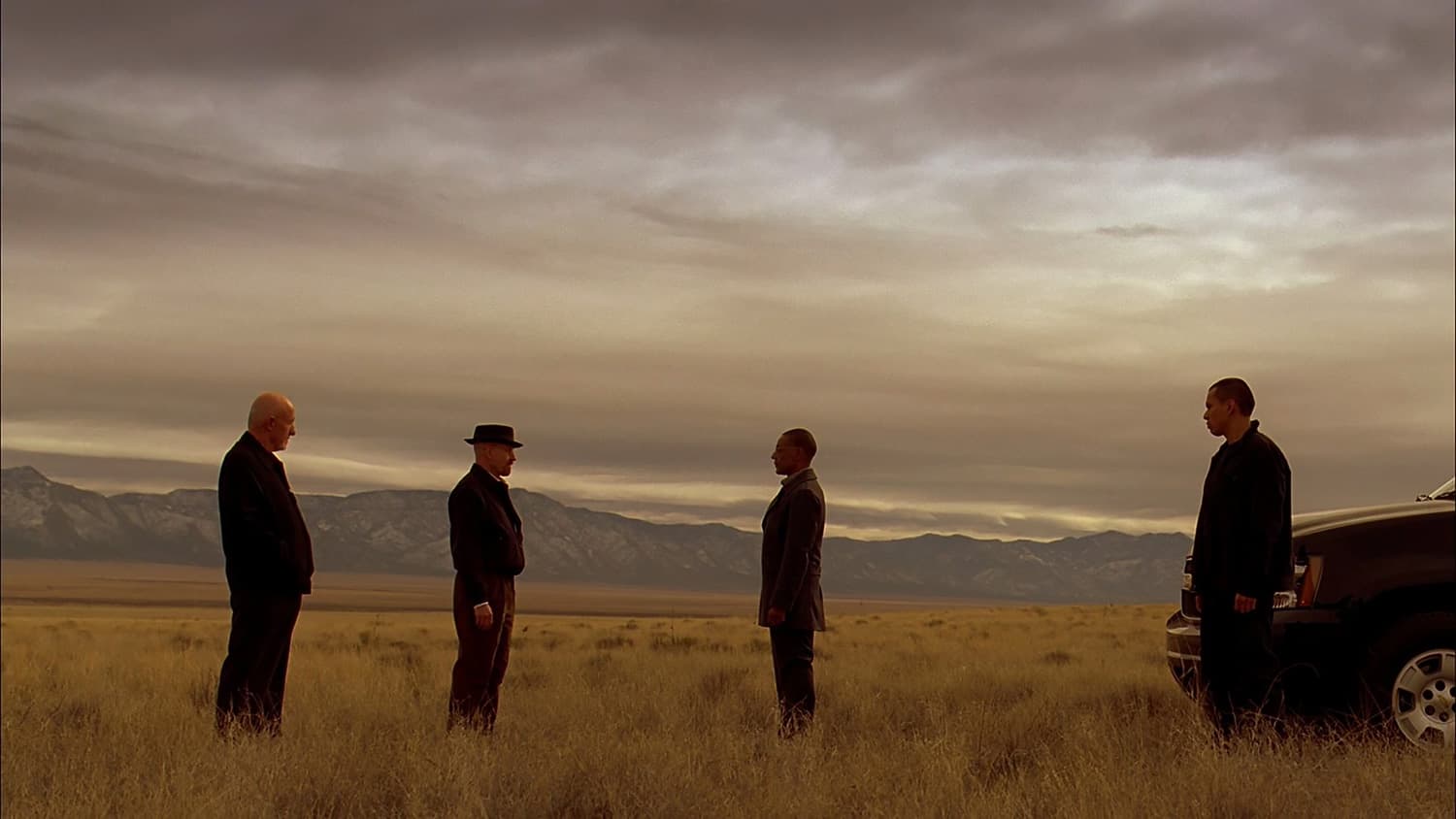 A wide-angle, sepia-toned cinematic shot from the TV series Breaking Bad featuring four men standing in an expansive, dry desert grassland.