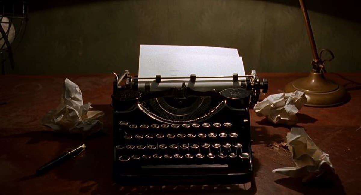 A close-up cinematic shot of a vintage black typewriter centered on a dark wooden desk under a warm lamp light. A blank white sheet of paper is loaded into the machine. Several crumpled balls of paper are scattered around the desk next to a fountain pen, against a dim, textured wallpaper background.