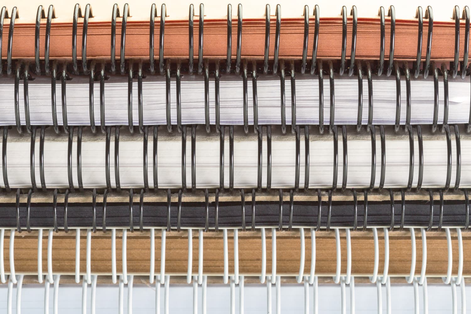 A close-up view of a stack of multiple spiral-bound notebooks and screenplays piled horizontally. The image highlights the repetitive texture of the black and white metal coils holding various brown and white pages together.