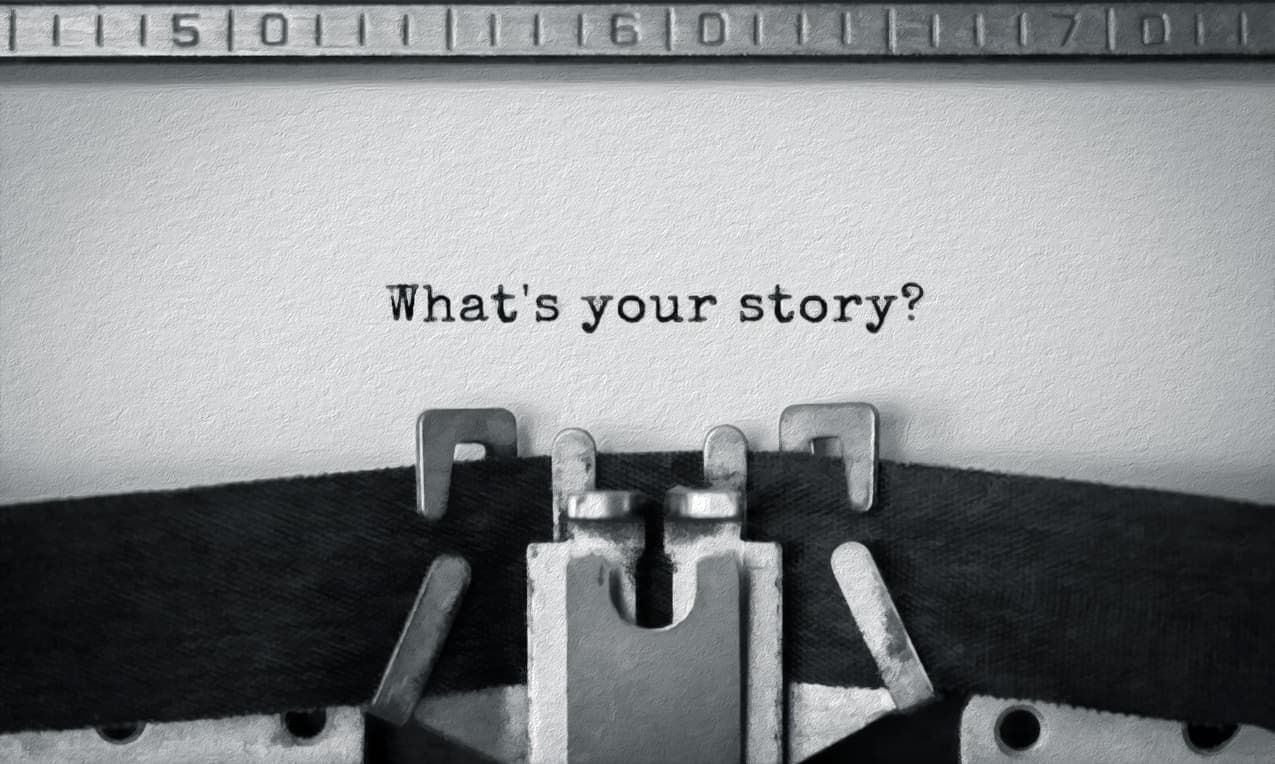 A black and white close-up of a vintage typewriter platen holding a sheet of textured paper. Centered on the paper is the typed text: "What's your story?" with the question mark. A numbered metal ruler is visible above the paper.